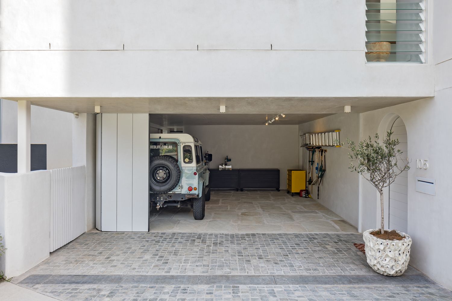 Open garage of a modern white house with a vintage off-road vehicle, paved floor and a potted olive tree.