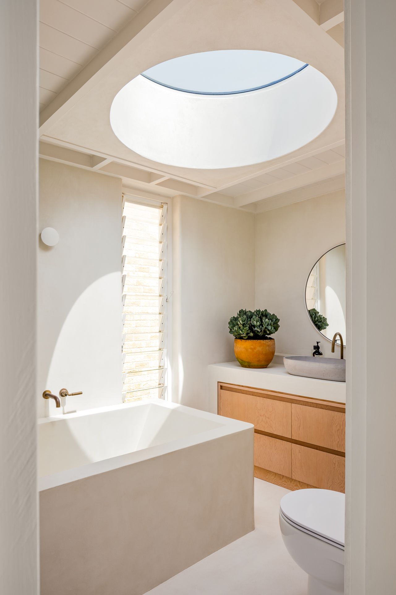 Minimalist bathroom with round skylight, plaster bathtub, brass taps, oak vanity, round mirror and potted plant in a bright space.