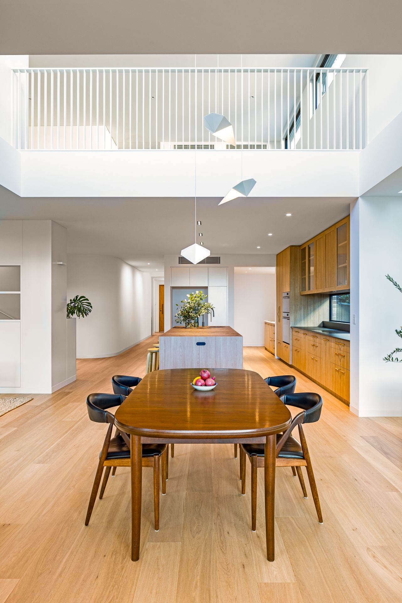 Airy open-plan dining area with oak table, black chairs, sculptural pendant lights, timber kitchen cabinets and a balustraded mezzanine.