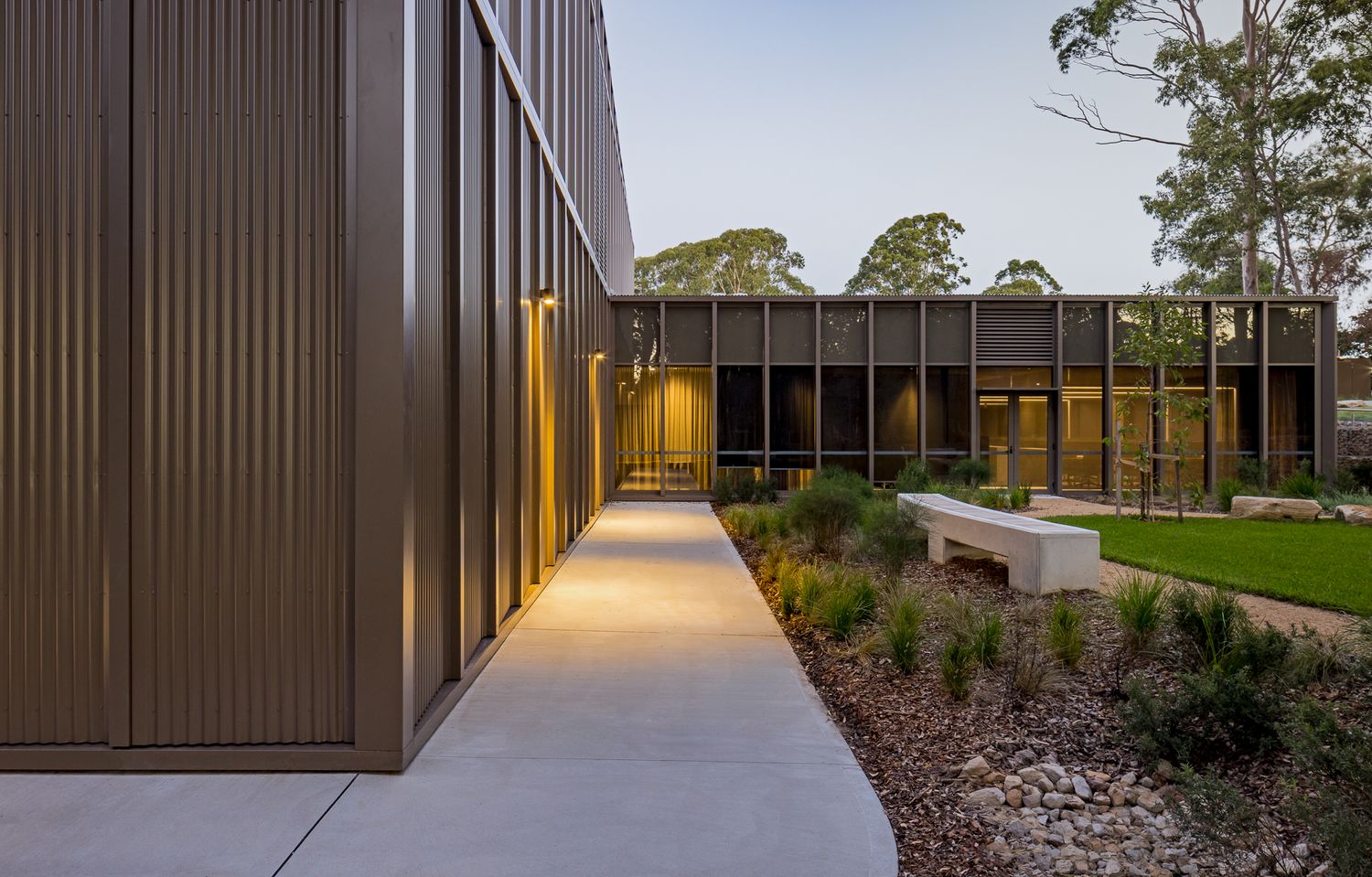 Concrete walkway lit by warm wall lights runs alongside corrugated bronze cladding toward a glazed courtyard with native planting at dusk.