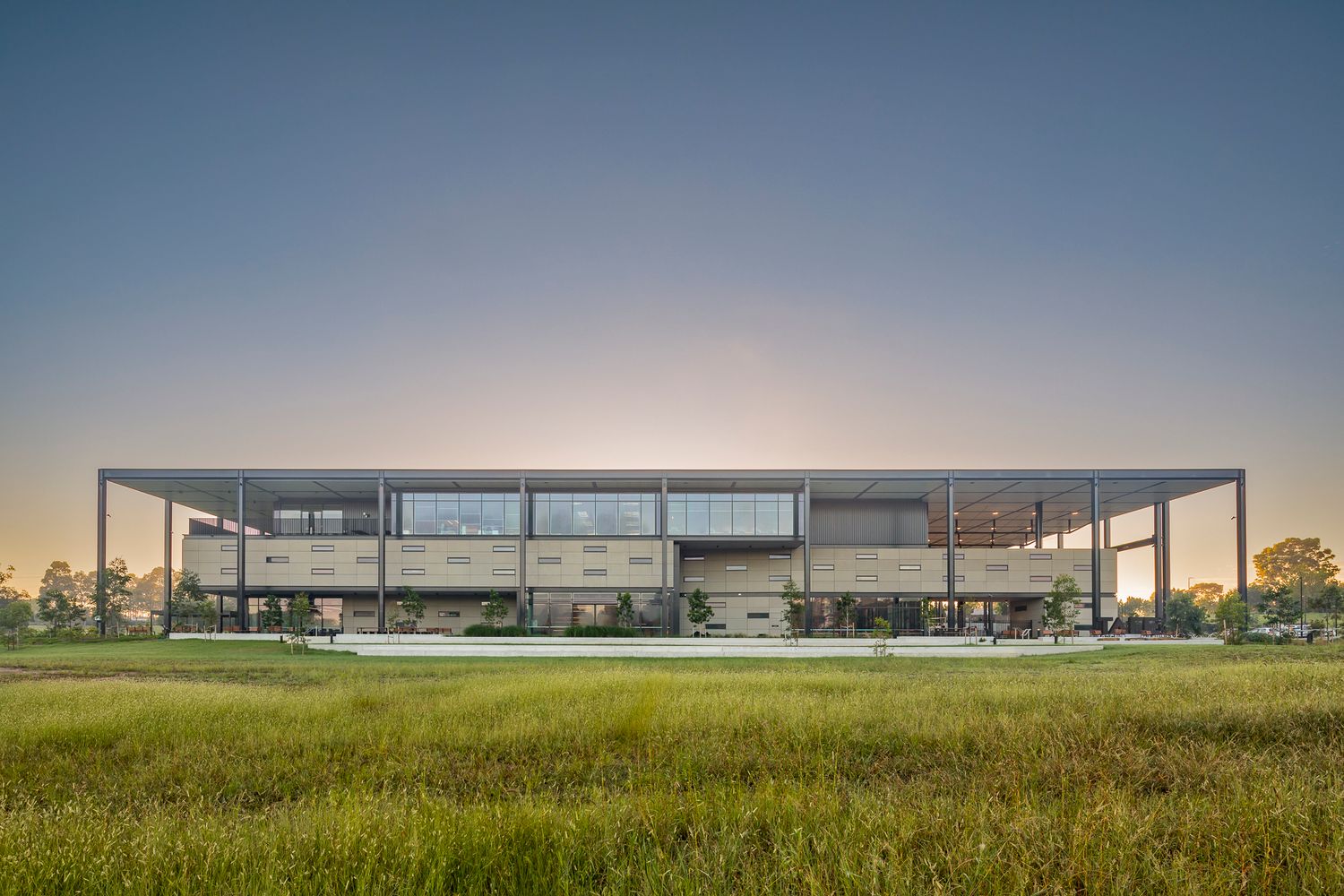 Symmetrical steel-framed modern building with glazed upper floor and wide overhanging roof across a grassy meadow at dusk.