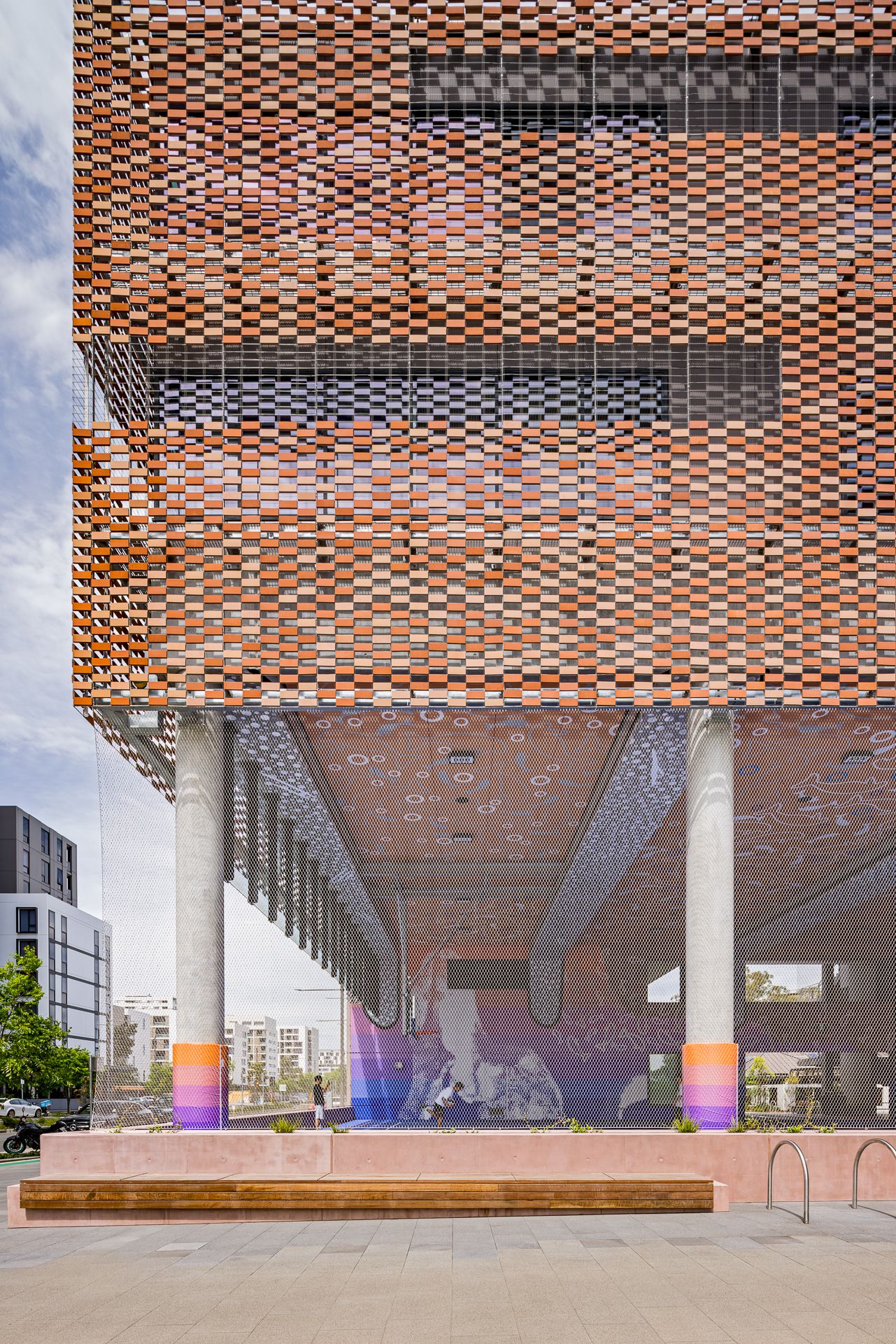 Modern building with a perforated brick facade elevated on concrete columns with colorful striped bases above a covered plaza with a mural.