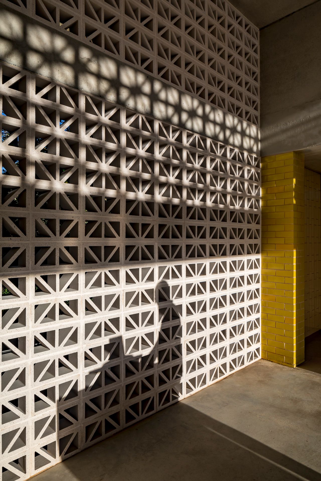 Sunlight casts patterned shadows through a white breeze block wall beside a bright yellow brick column and a polished concrete floor.