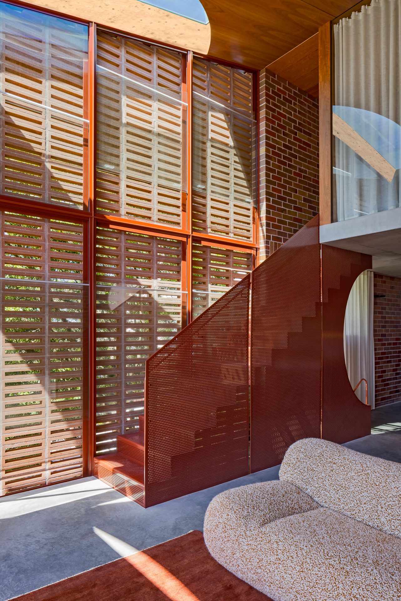 Interior double-height space with timber louvre screens, a red perforated metal staircase, brick wall and a bouclé sofa.