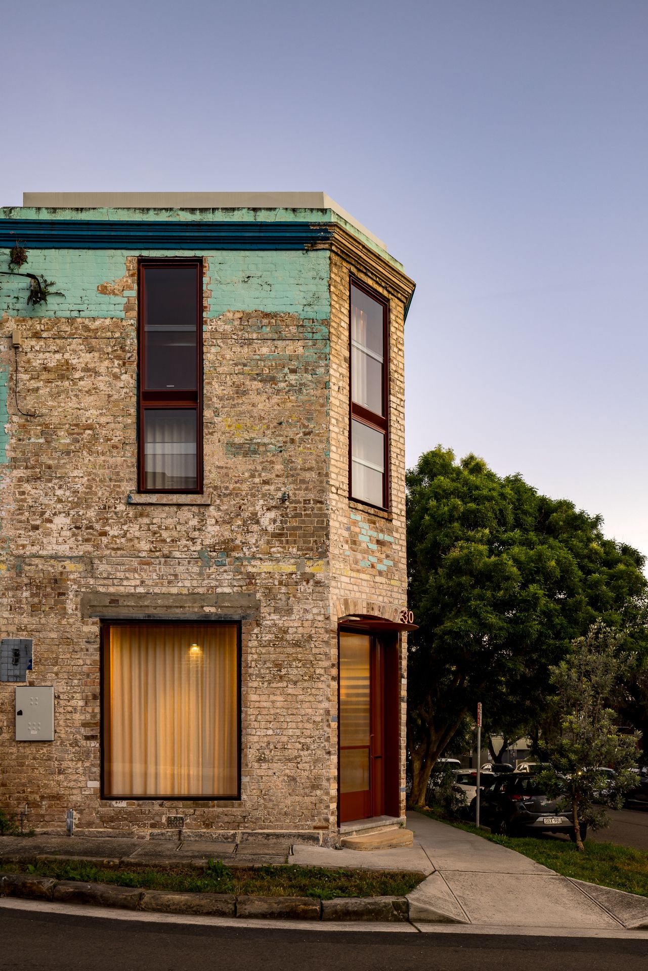Restored heritage brick corner building with weathered teal paint, red-framed timber windows and a tree in the evening light.