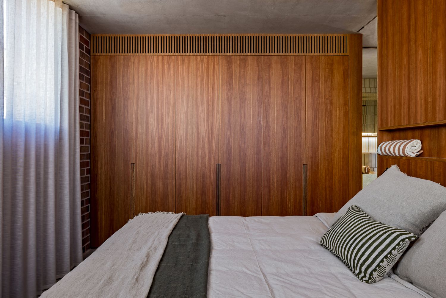 Modern bedroom with floor-to-ceiling timber wardrobes, a white linen bed with a gray throw and a striped cushion beside a sheer curtain.