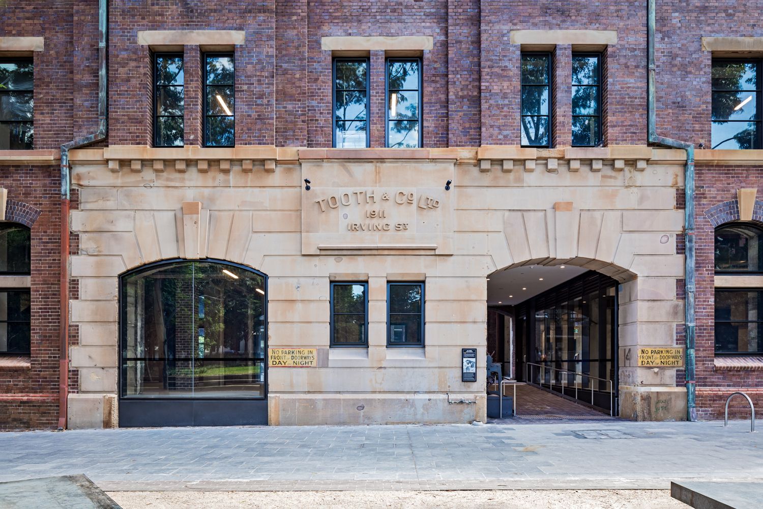 Heritage sandstone and red brick building entrance with arched glass doors and an engraved stone sign above a paved plaza.