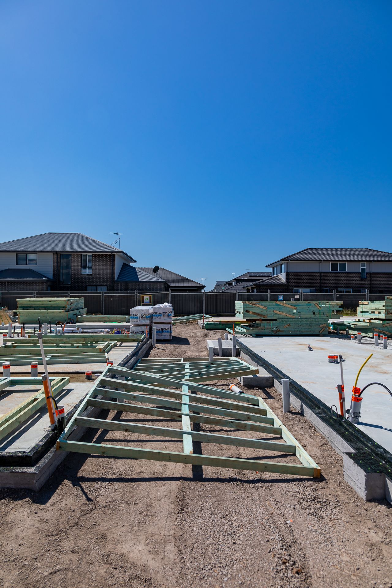 Residential construction site with timber floor framing, stacked building materials and completed houses in the background.