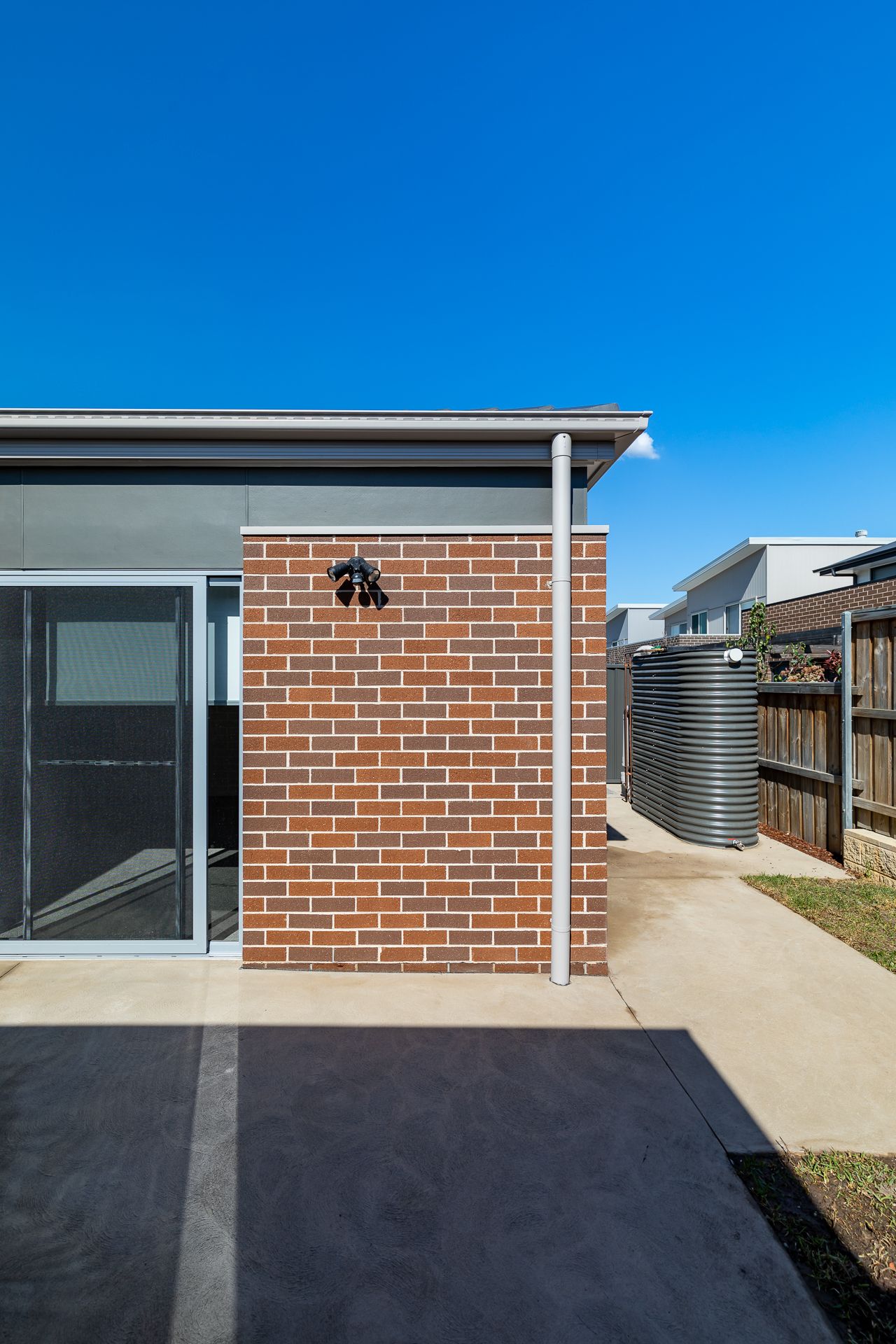 Red brick exterior wall of a new home with a white downpipe, sliding screen door and a corrugated water tank beside the fence.