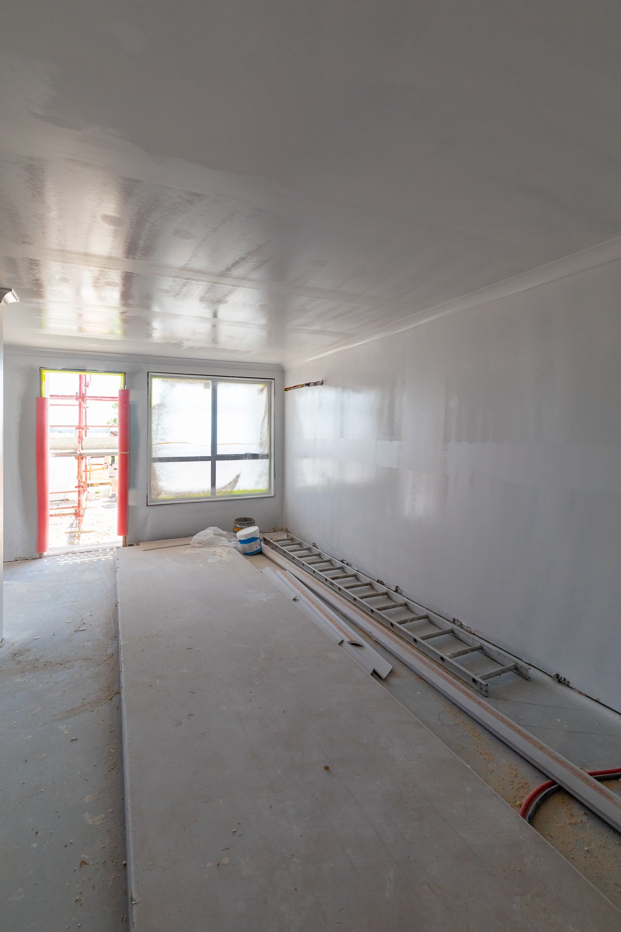 Unfinished interior room with plasterboard walls, a ladder on the floor and a window and doorway under construction.