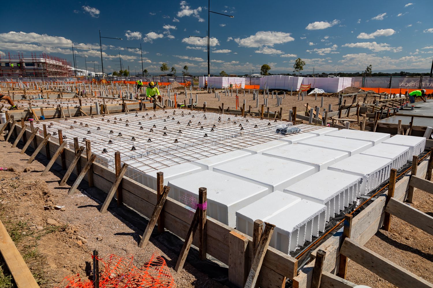 Rows of white polystyrene waffle pods laid on a house slab foundation with timber formwork and workers on a construction site.