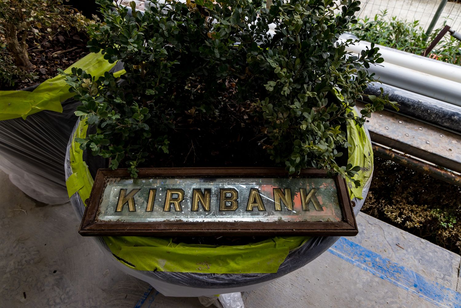 Wooden Kirnbank name sign resting on a planter of green shrubs beside yellow tape at a renovation site.