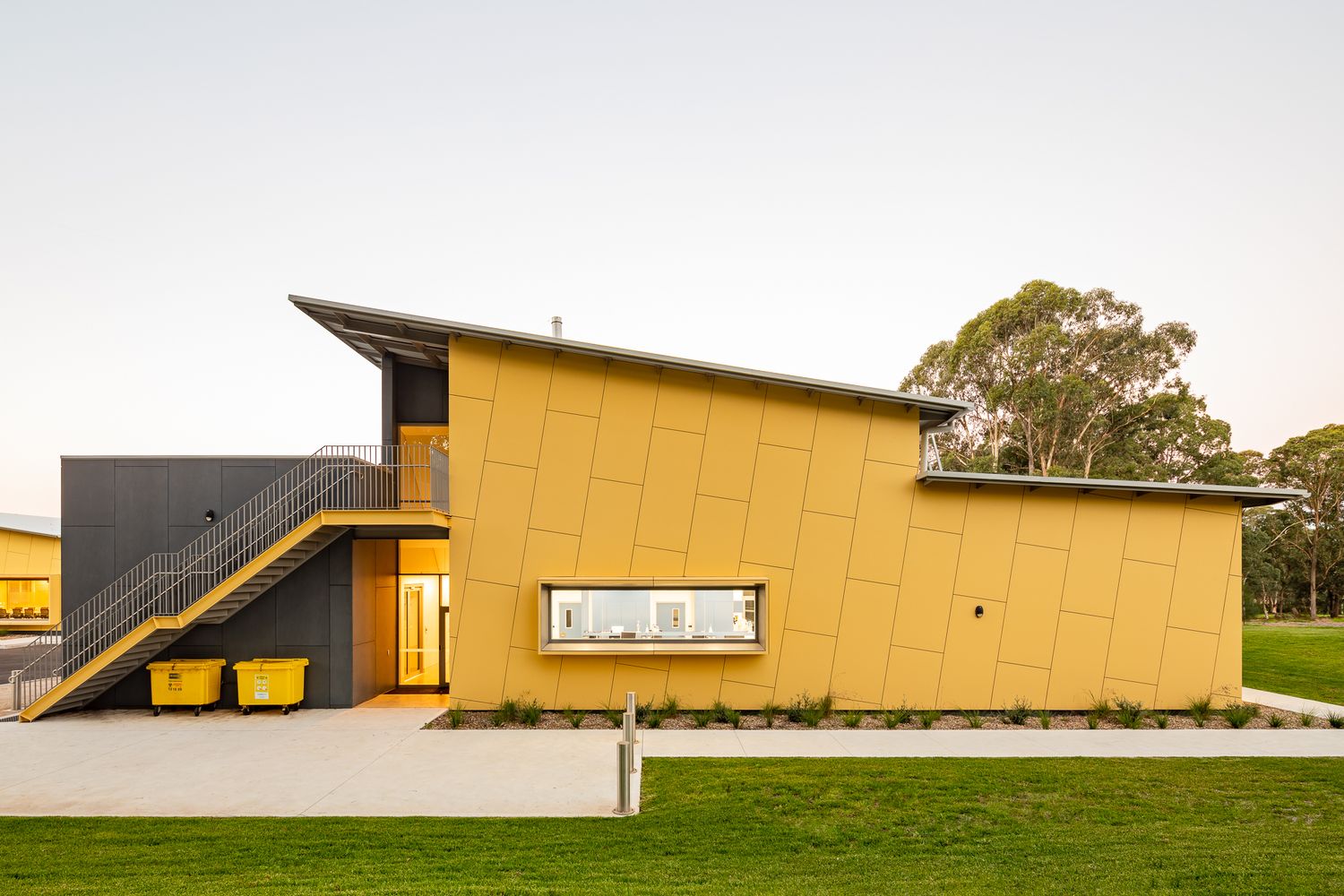 Contemporary yellow-clad building with a skillion roof, exterior staircase and two yellow bins set beside a lawn at dusk.