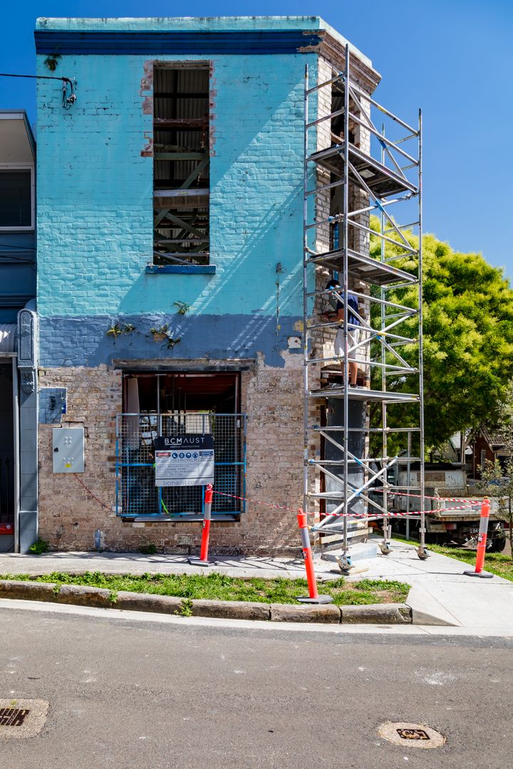 Blue-painted brick building with exposed brickwork and scaffolding tower under renovation beside a street with orange safety bollards.
