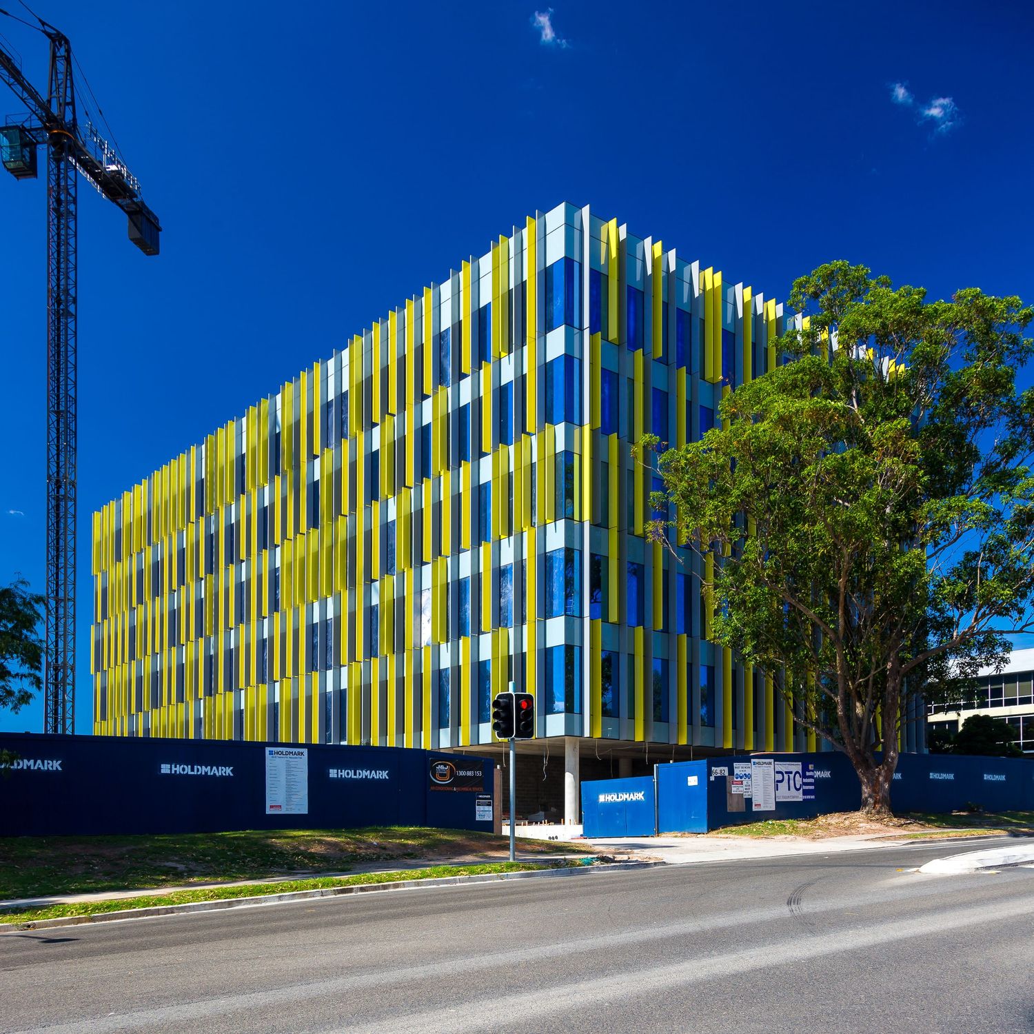 Modern multi-storey building with yellow, green and blue vertical facade under construction beside a crane and tree.