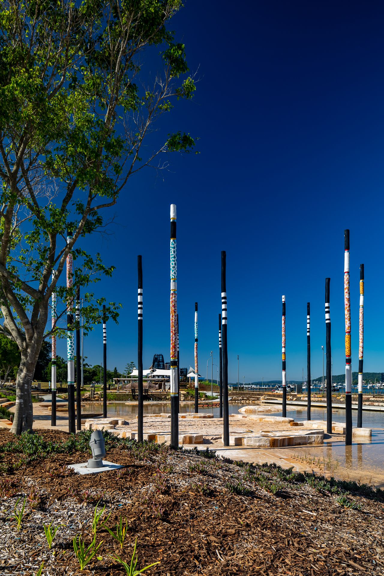 Colorfully painted tall poles rise beside a tree and sandstone water play area under a deep blue sky.