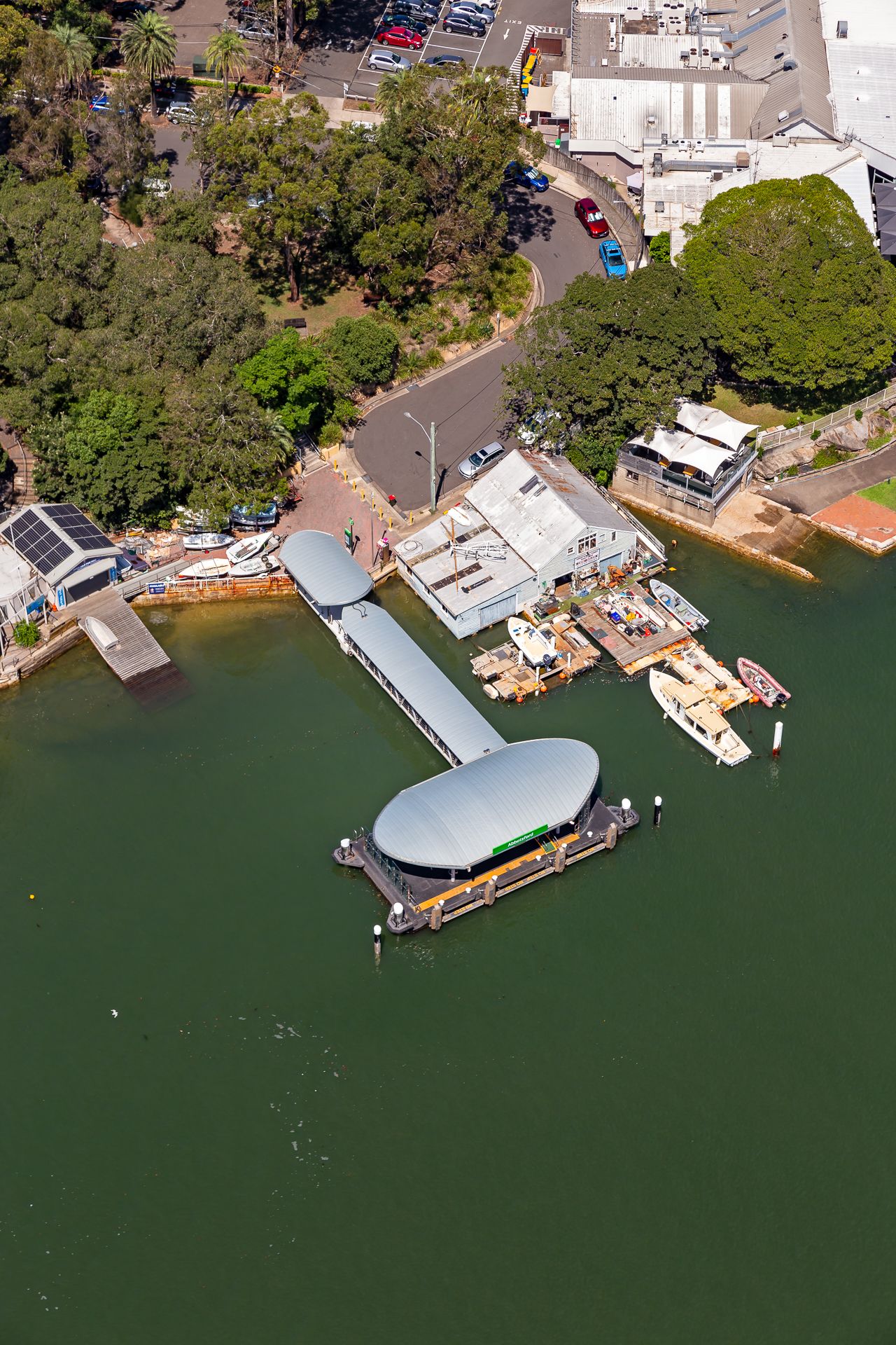 Aerial view of a waterfront ferry wharf with a curved-roof pontoon, jetty, moored boats, and harbourside buildings among green trees.