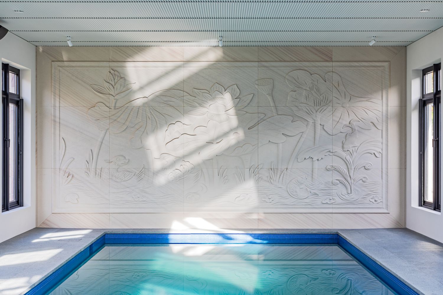 Indoor swimming pool with turquoise water and a large white carved relief mural of botanical designs lit by sunlight from tall windows.