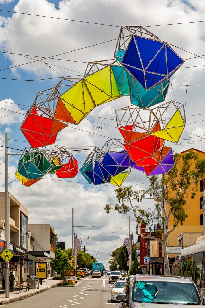 Colorful geometric lantern sculptures arch over a suburban shopping street under a blue sky with clouds.