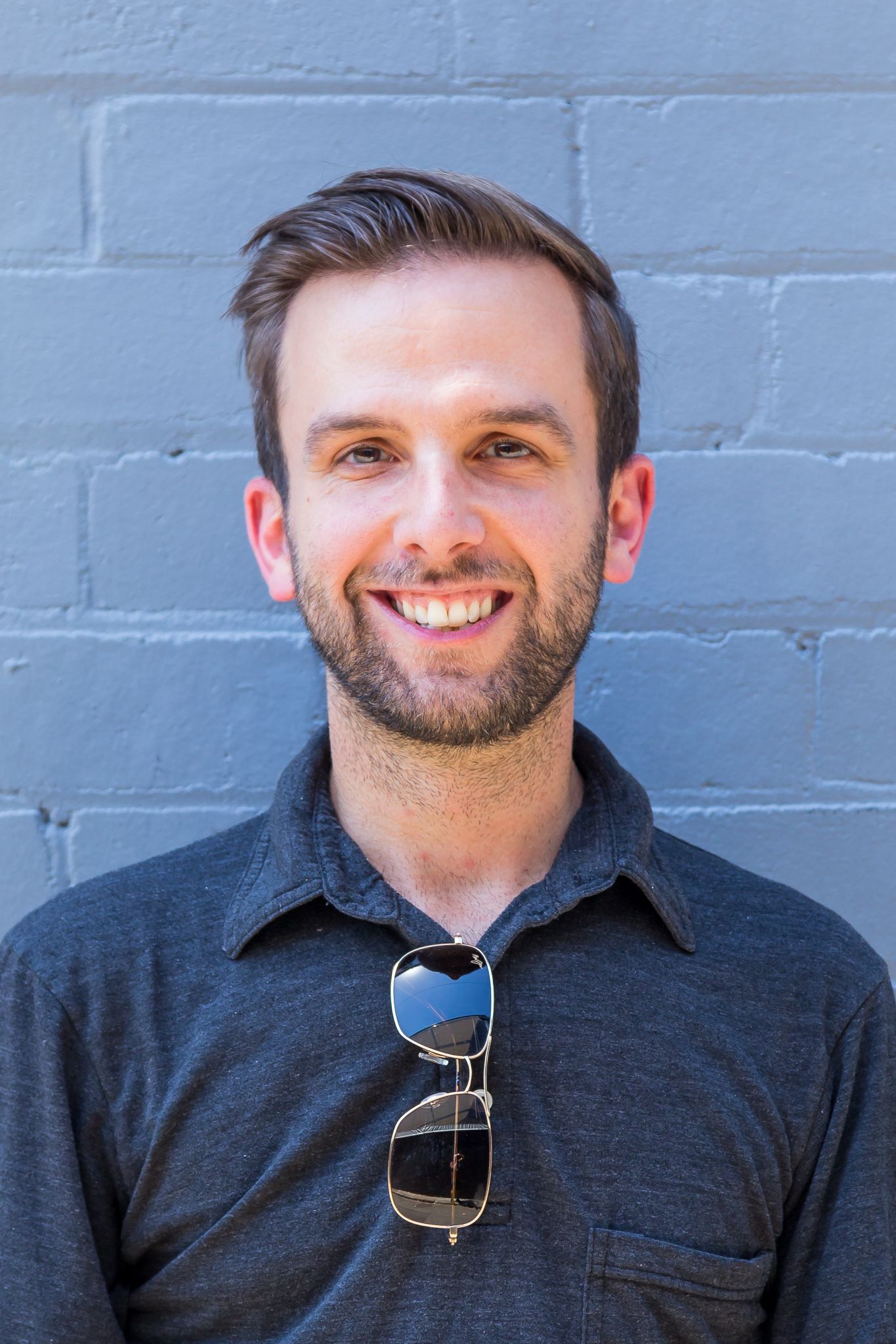 Smiling man with short brown hair and a beard wears a gray collared shirt with sunglasses hanging from the collar against a blue brick wall.