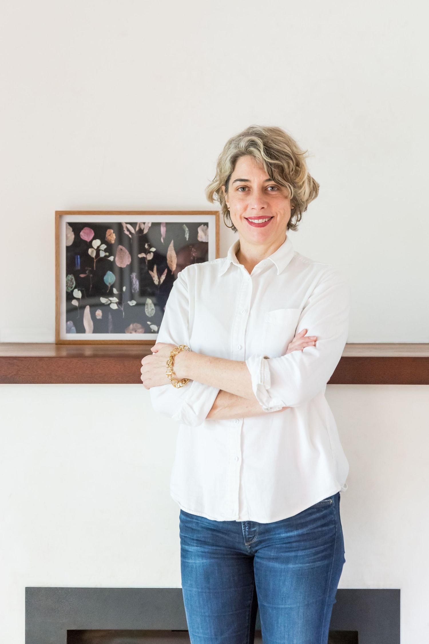 Smiling woman with short curly hair and a white shirt stands with arms crossed beside a framed floral artwork on a wooden mantel.