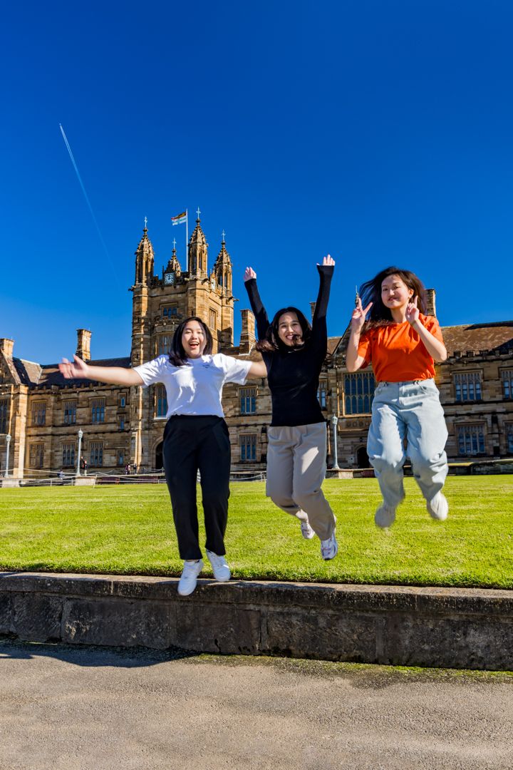 Three joyful students jump on a sunny lawn in front of a historic sandstone university building under a blue sky.