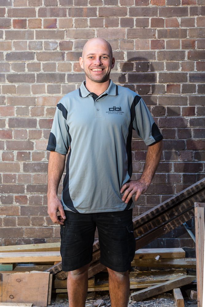 Smiling bald man in a gray polo shirt and black shorts stands in front of a brick wall beside stacked timber on a worksite.