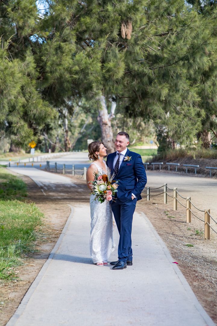 A bride and groom in a navy suit share a happy moment holding a bouquet on a path lined with trees.