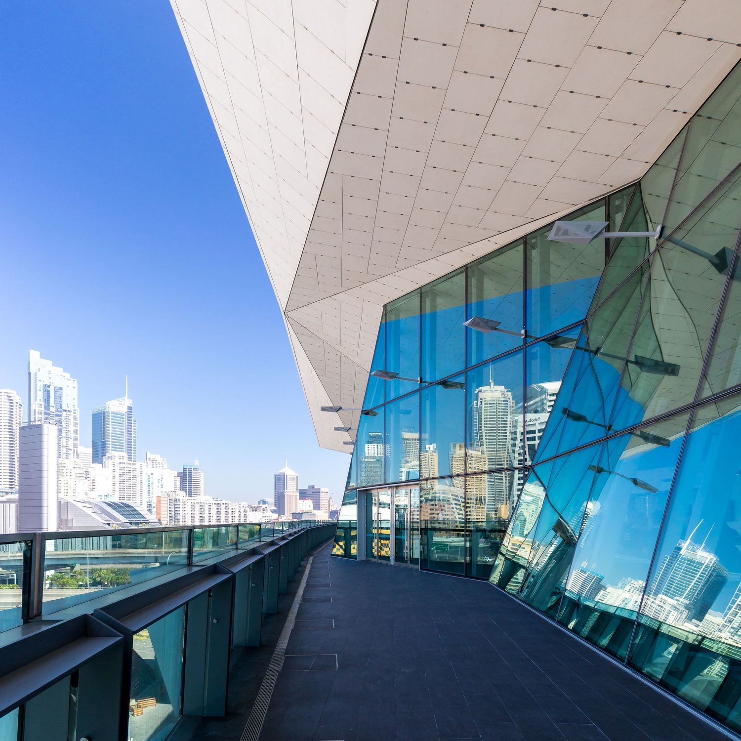 Outdoor walkway beside a slanted glass facade and angular ceiling overlooking the city skyline on a sunny day.