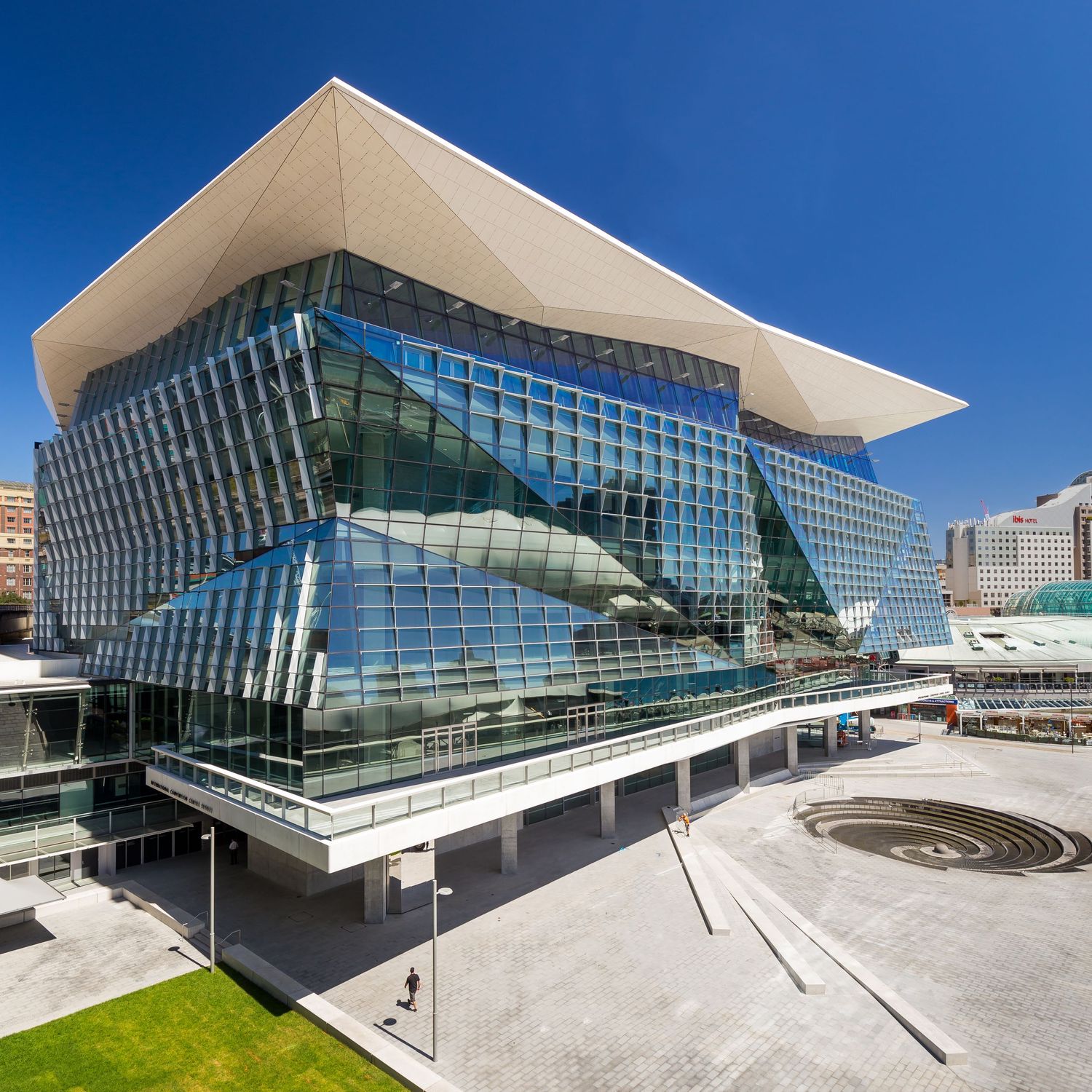 Angular glass-faced convention centre with a sweeping white cantilevered roof and a paved public plaza under a clear blue sky.