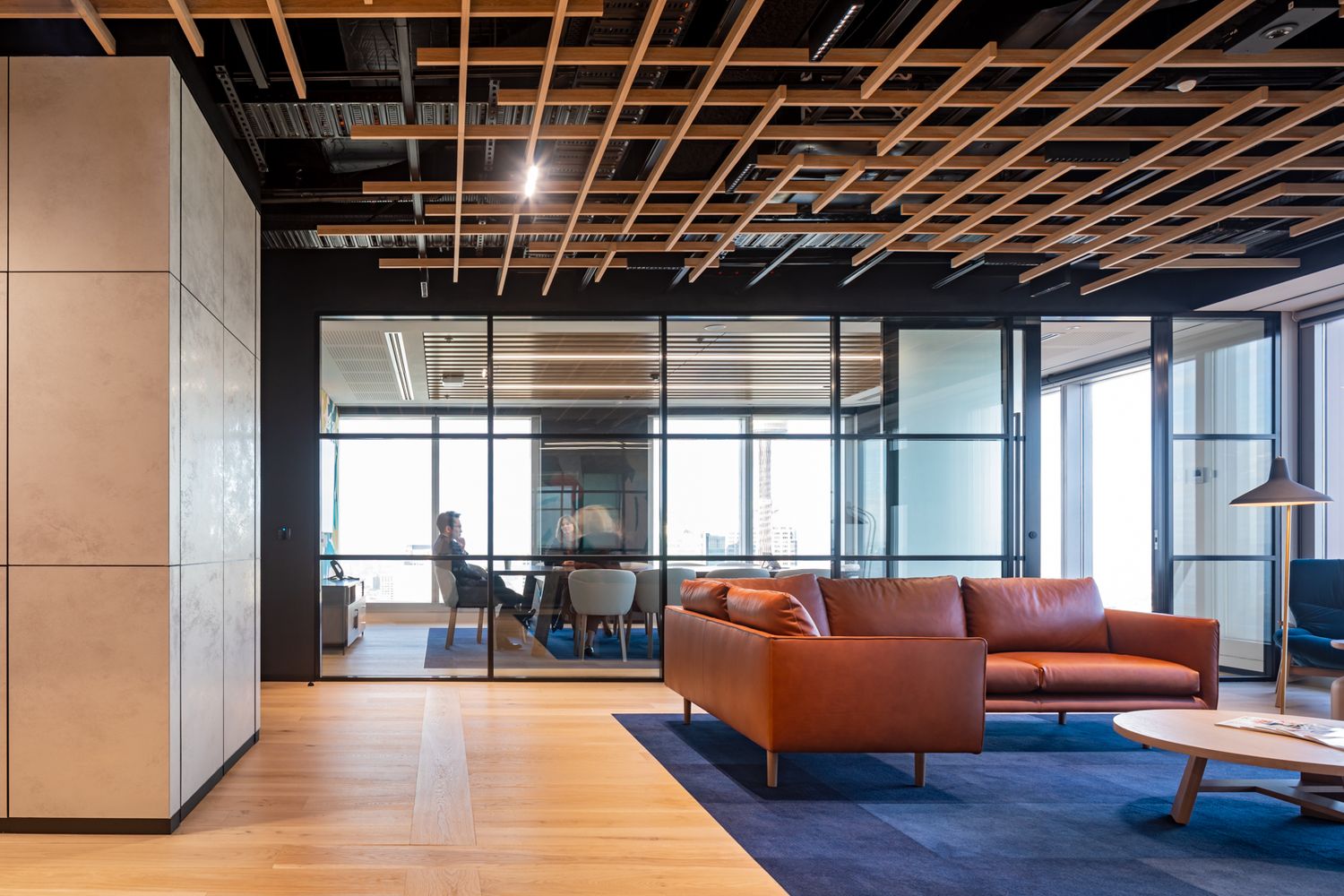 Modern office lounge with tan leather sofa, blue carpet, timber slat ceiling feature and glass-walled meeting room in the background.