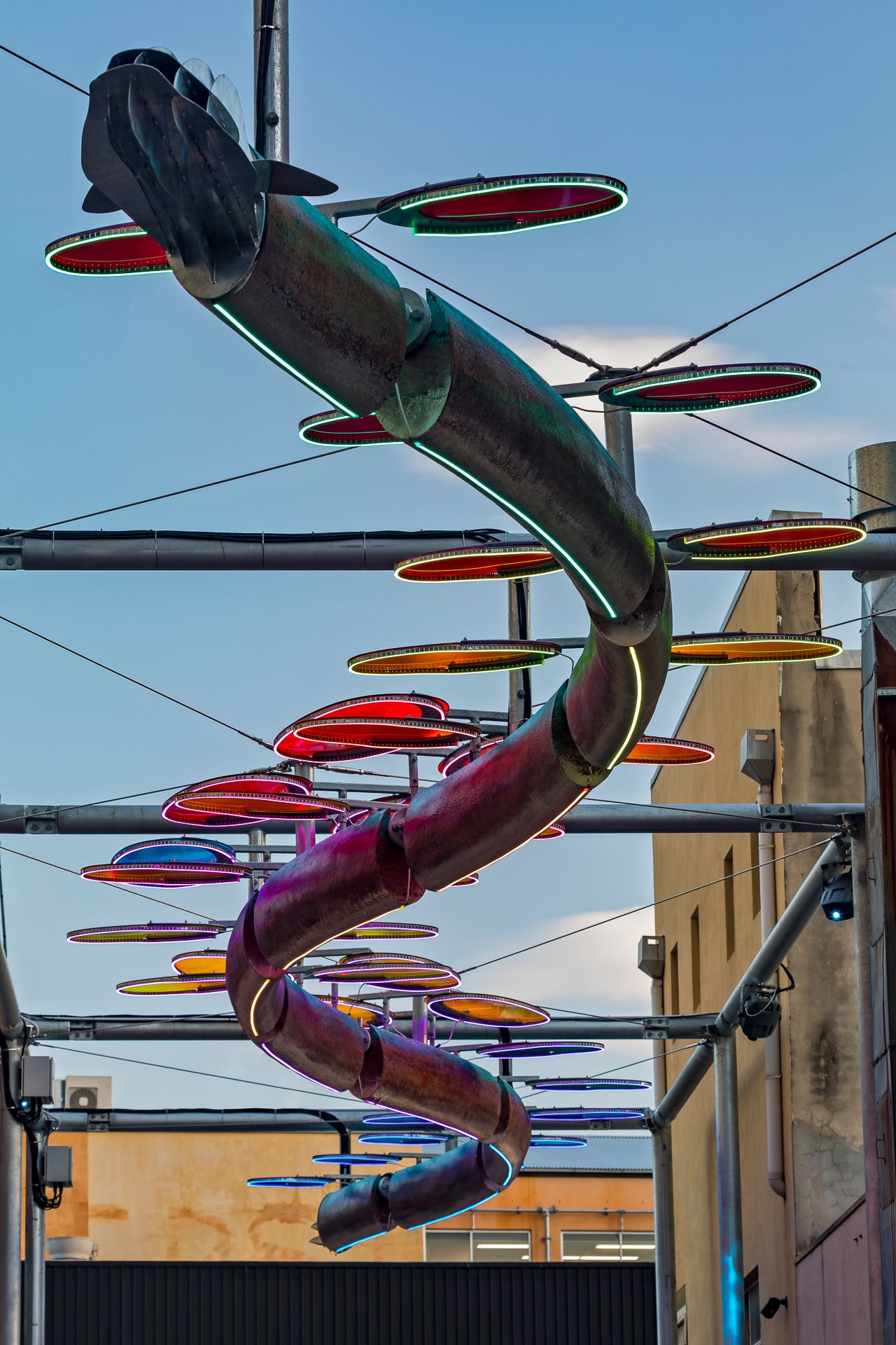 Fish-headed serpent sculpture with neon-edged colorful oval discs hanging from steel frames above an urban laneway.