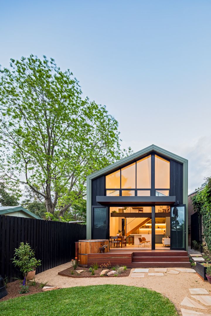 Gabled modern home with a black timber facade, large glazed windows and a glowing interior, set in a garden with a tall tree.