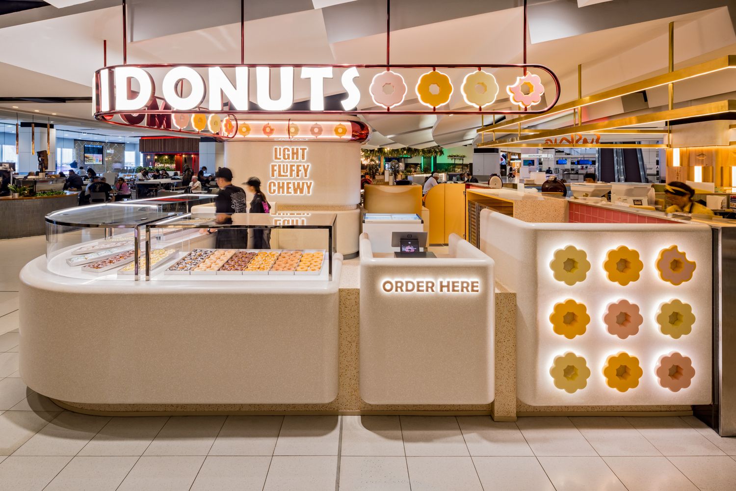 Bright I Donuts shop in a shopping centre with a glowing sign, donut display counter, order here station and flower-patterned wall.