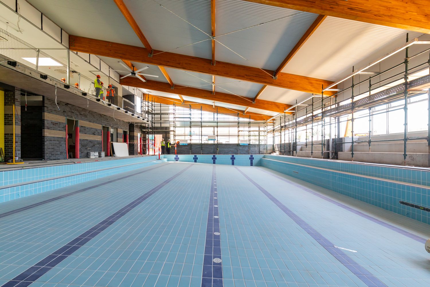 Indoor swimming pool hall with light blue tiled pool, lane markings, exposed wooden roof beams, and large side windows.