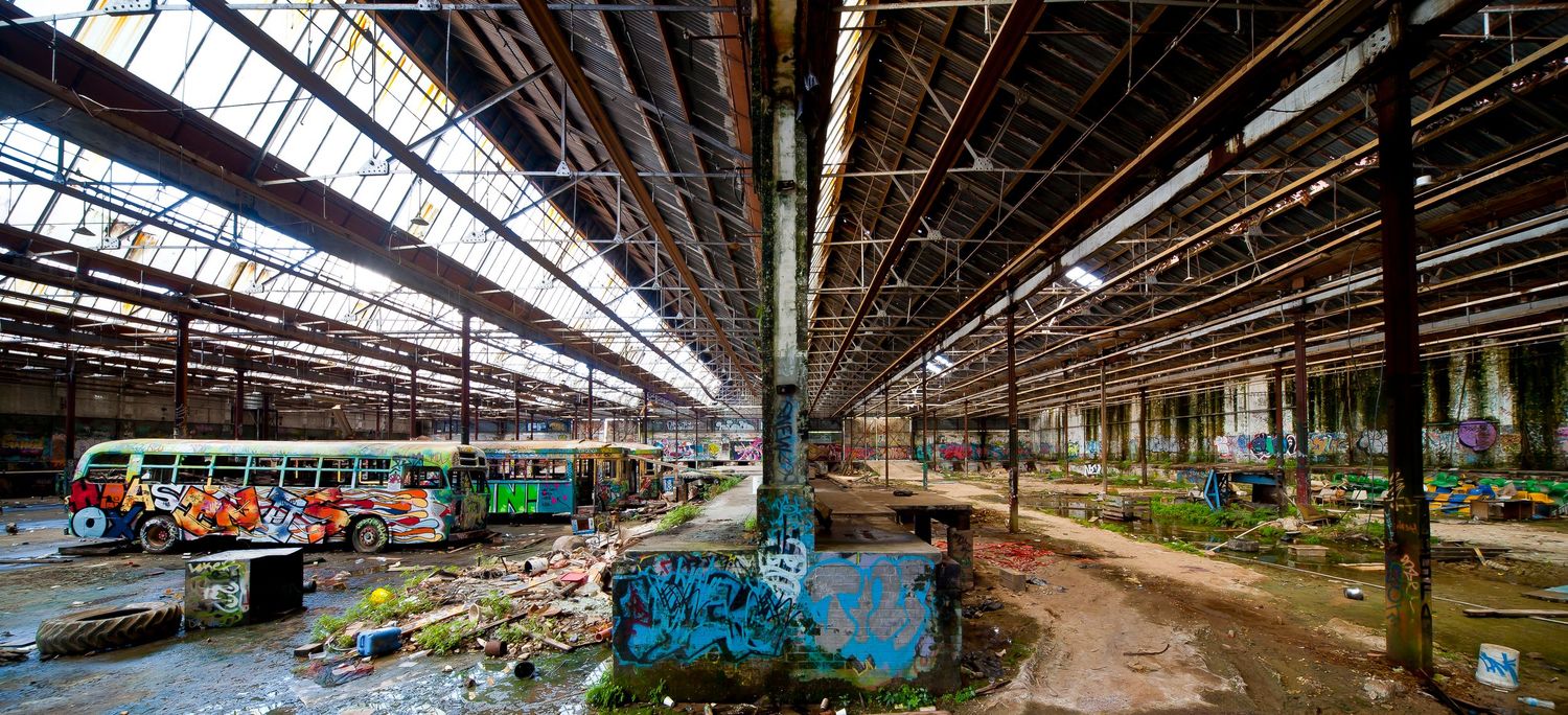 Vast abandoned industrial hall with steel roof trusses, graffiti-covered columns and a derelict colorful bus inside.