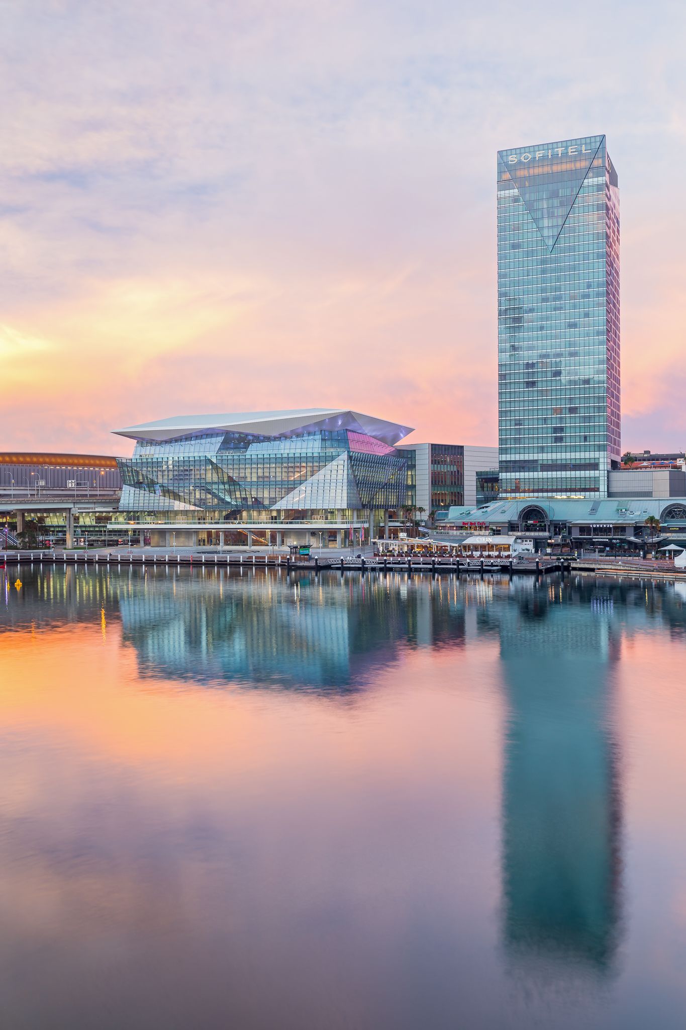 Sofitel tower and a glass-fronted waterfront building reflect in calm harbor water at sunset under a soft pink and orange sky.