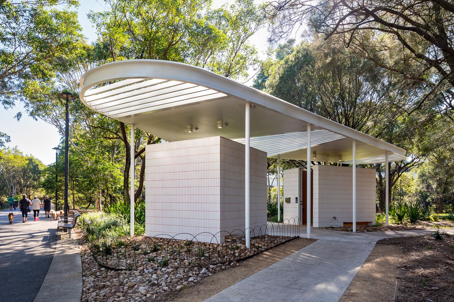 Curved white roof pavilion with tiled walls and slender columns beside a park path, with pedestrians walking in the distance.