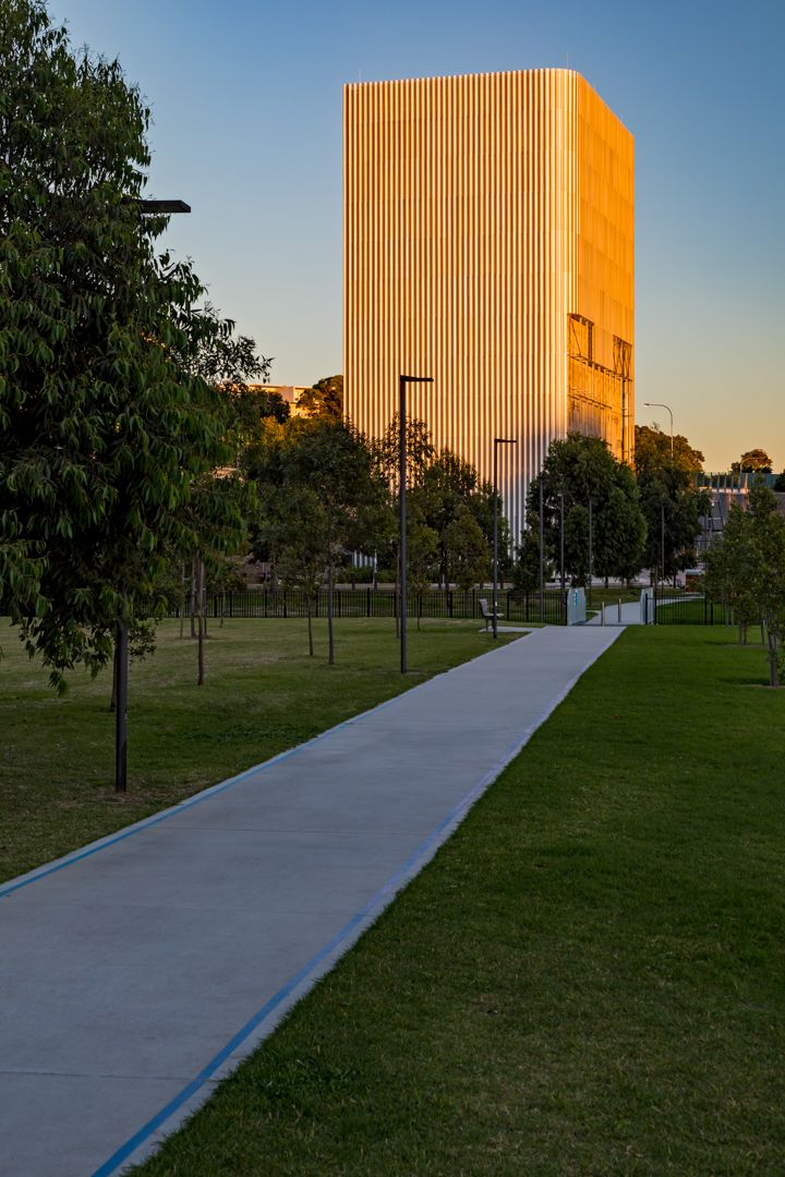 Tall rectangular tower with golden vertical fins glowing in sunset light, seen from a green park with a concrete pathway and trees.