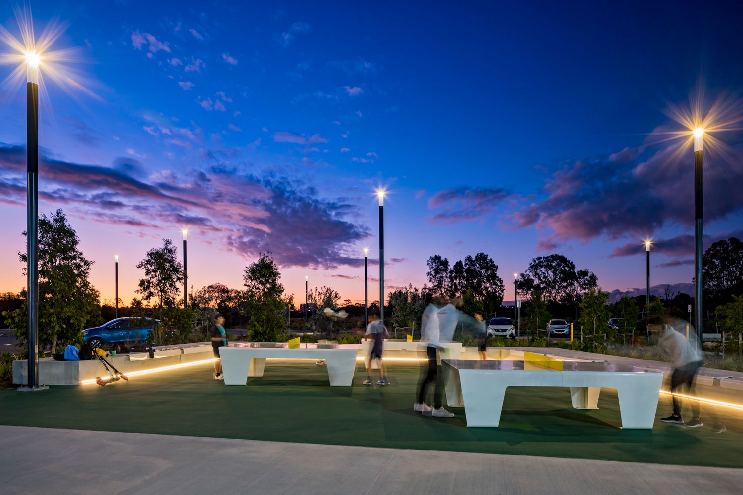 Illuminated park at dusk with tall lamp posts, modern white benches and people walking beneath a purple and blue twilight sky.