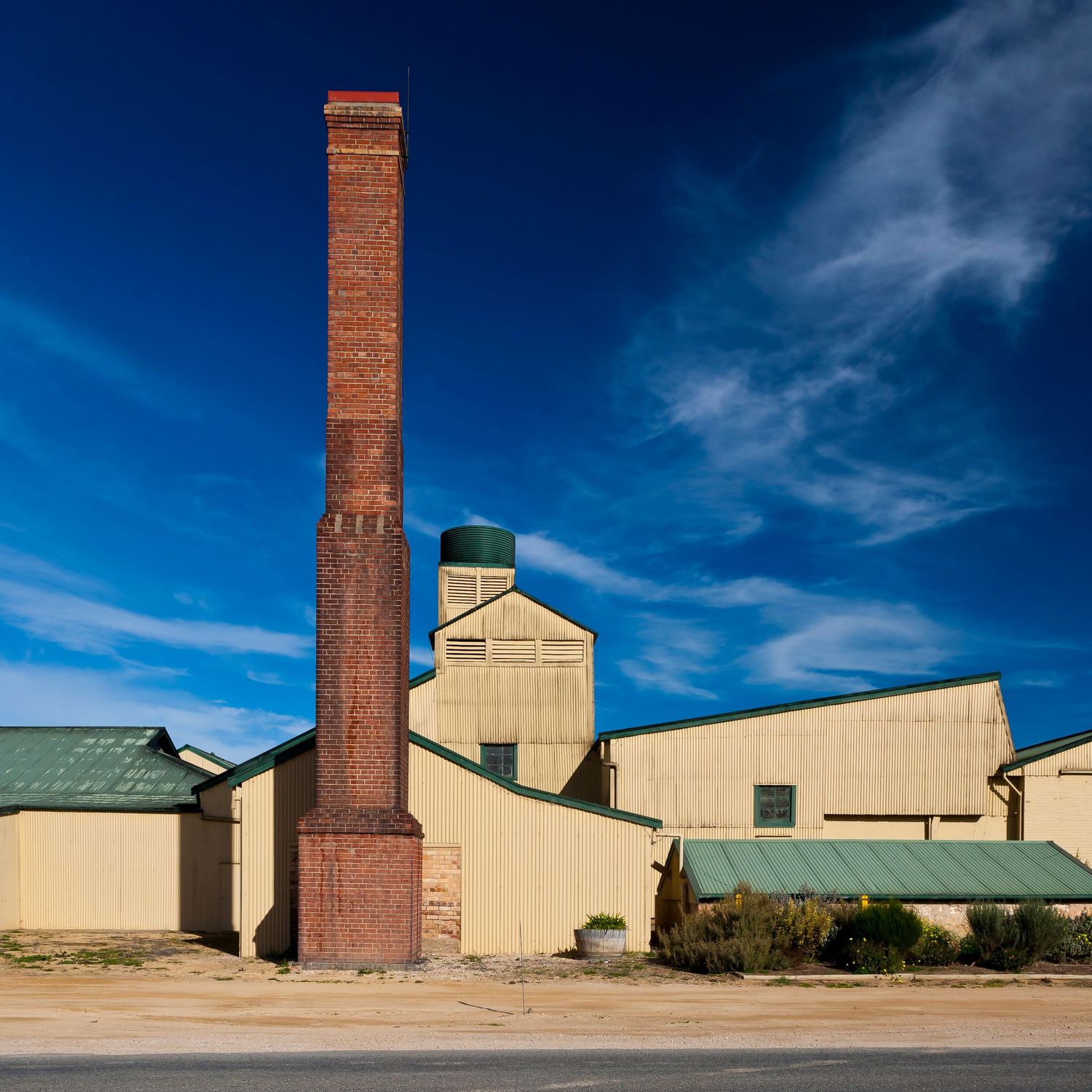 Tall red brick chimney beside cream industrial sheds with green roofs and a small water tank under a deep blue sky.