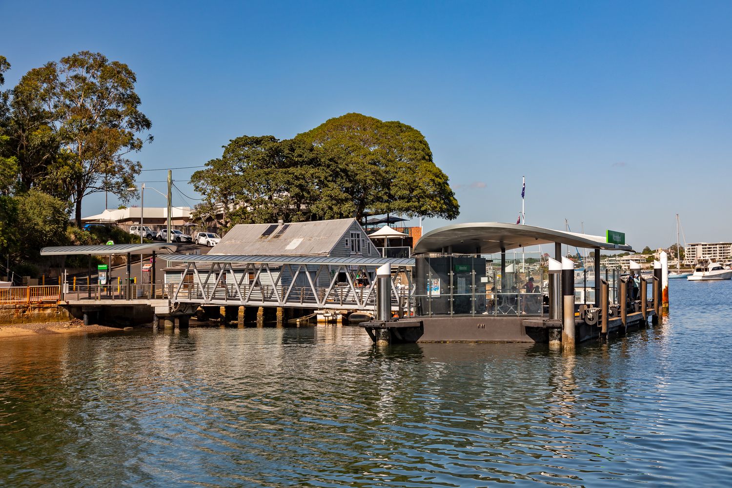 Waterfront ferry wharf with covered jetty and pontoon beside leafy green trees on a sunny day over calm blue harbour water.