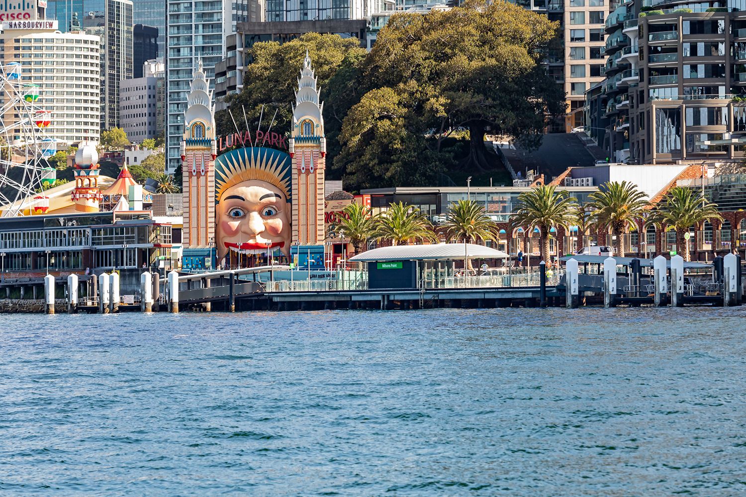 Harbourside ferry wharf near the giant smiling face entrance of Luna Park Sydney, viewed from the water across the blue harbour.