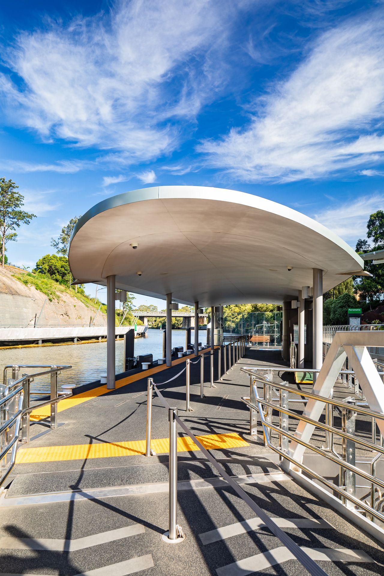 Modern curved-roof ferry wharf with tactile yellow paving and steel railings under a bright blue sky with wispy clouds.