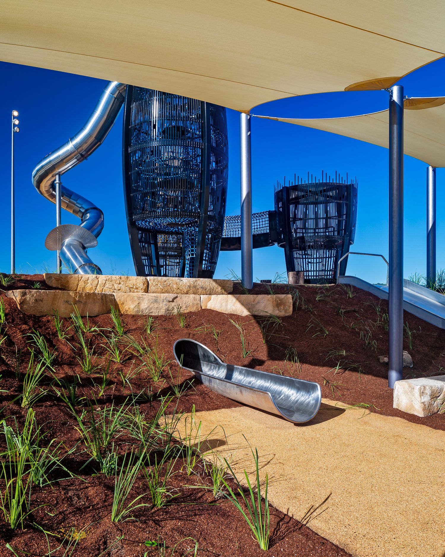 A silver slide curves down a landscaped slope beneath a shade sail, with a black lattice play tower behind.