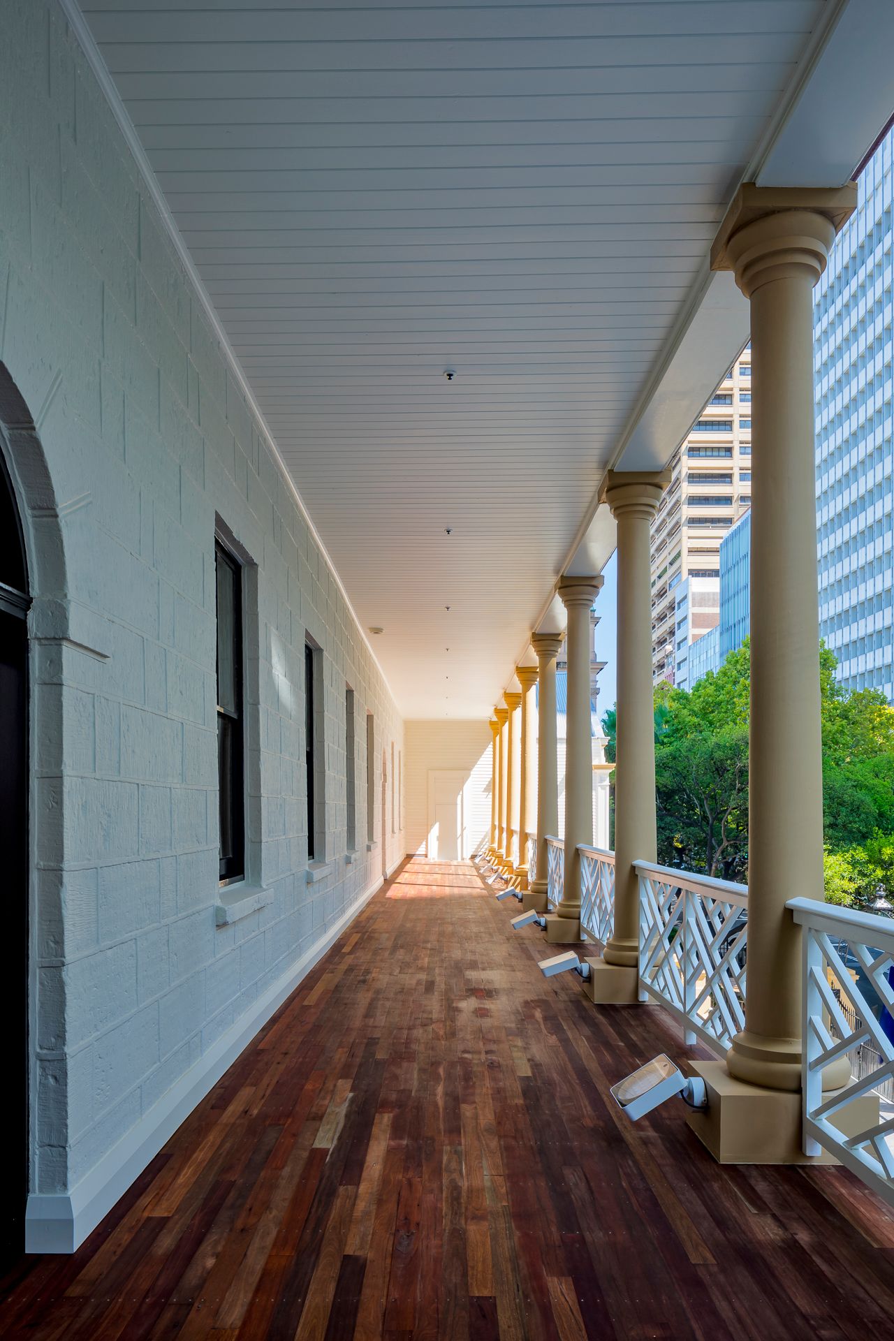 Long covered timber-floored veranda with cream columns and lattice railings beside a white stone wall, overlooking city high-rise buildings.