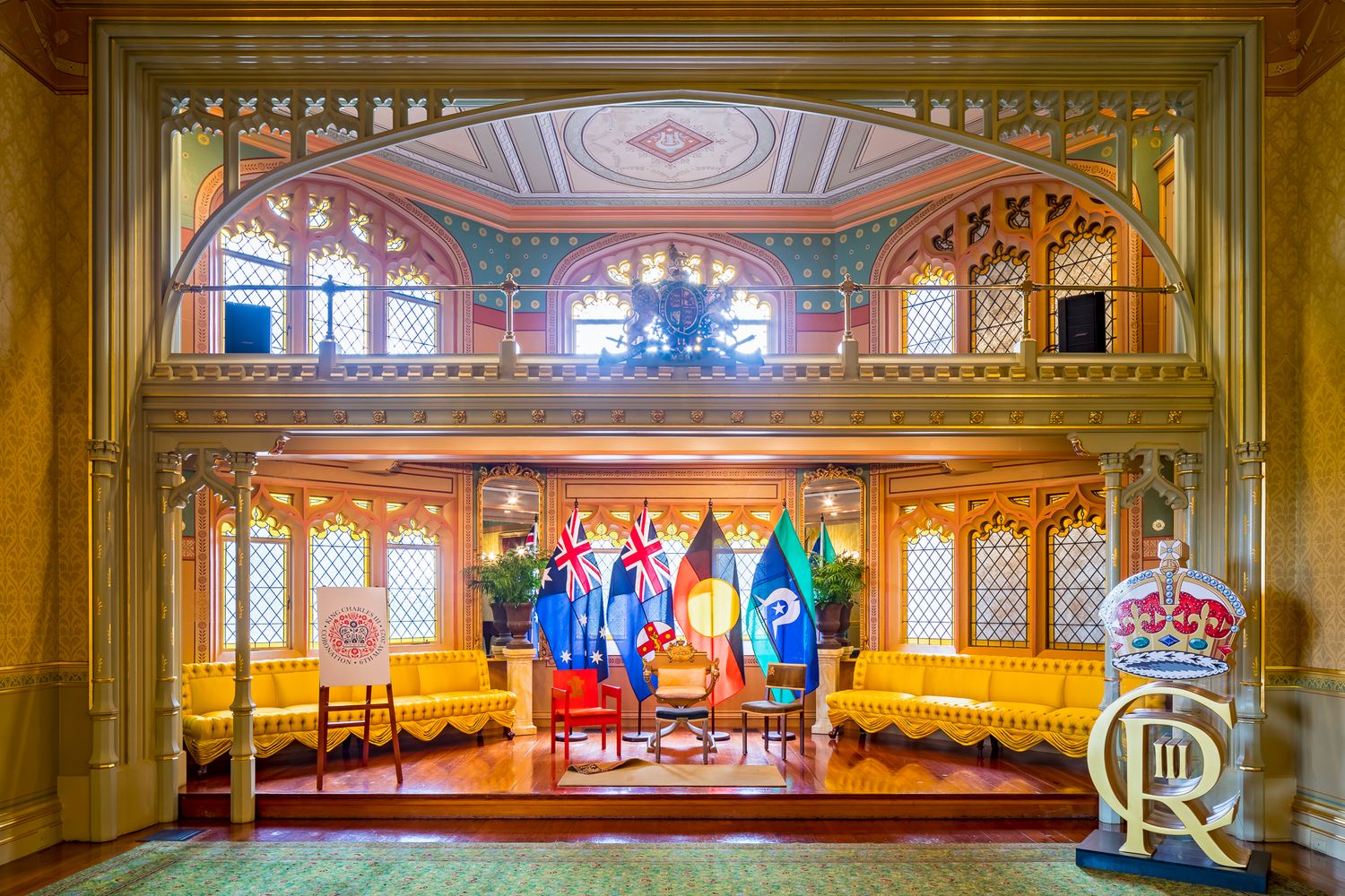 Elegant heritage hall with arched balcony, gold sofas, flags and a royal cypher on a stand beneath intricate trim.