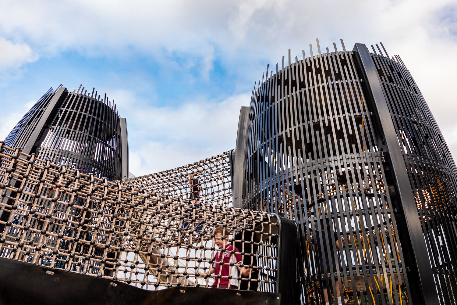 Tall black steel lattice climbing towers linked by a rope net bridge, with children visible against a blue and cloudy sky.