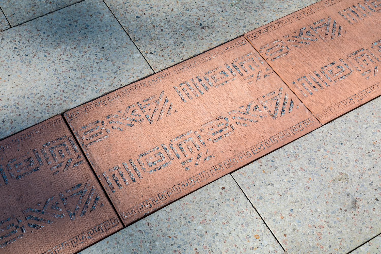 Close-up of terracotta-colored paving strips with engraved geometric patterns set into pale grey pavers in a public square.