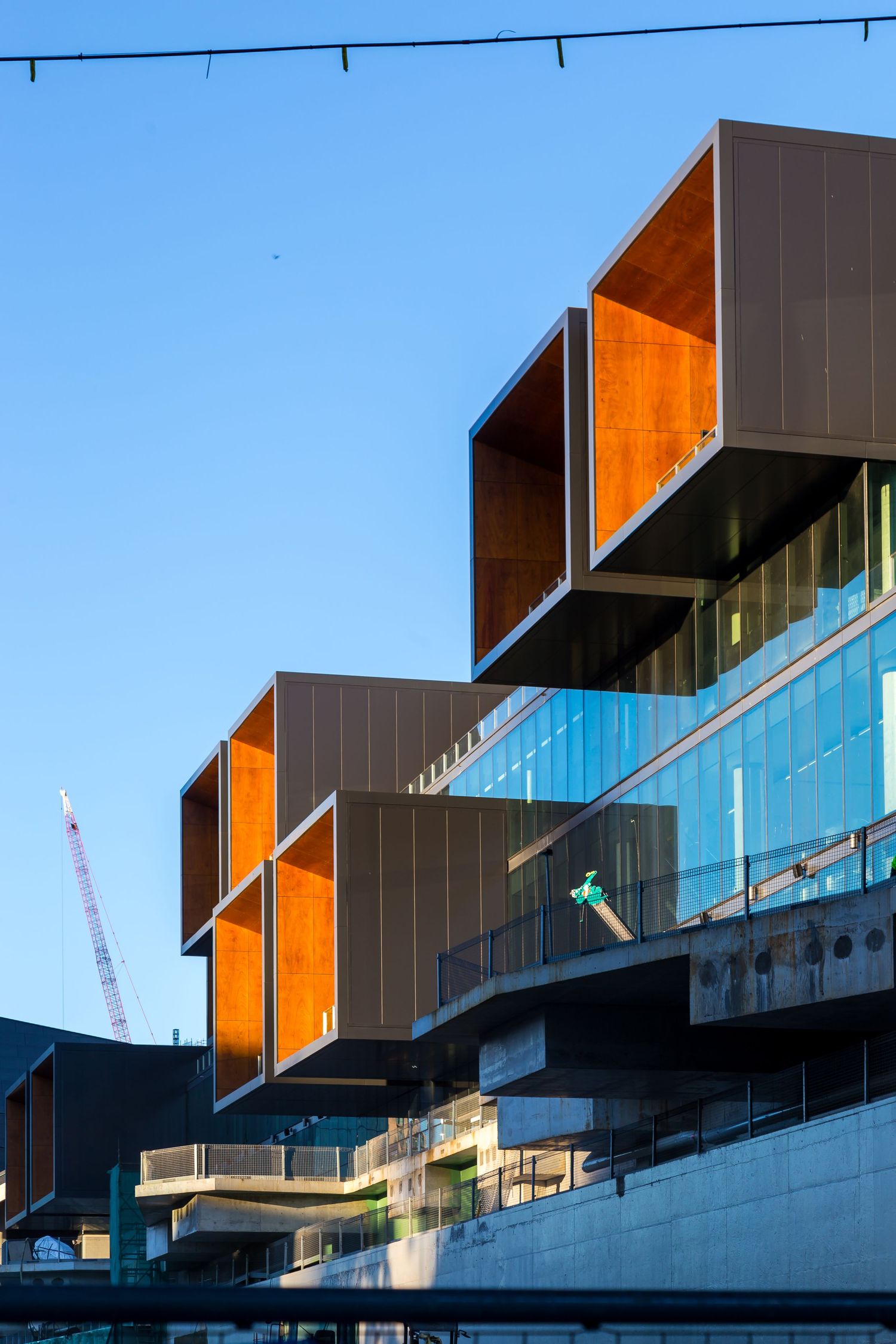 Stacked cantilevered boxes with orange timber interiors projecting from a modern glass building against a blue evening sky.